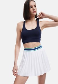 Granatowy crop top z szerokimi ramiączkami w połączeniu z białą plisowaną spódnicą z wielokolorowym paskiem w poziome pasy. Gładki materiał, sportowy fason.