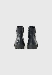 Bottines en cuir noir avec des semelles épaisses et des boucles latérales, vues de l'arrière sur fond blanc.