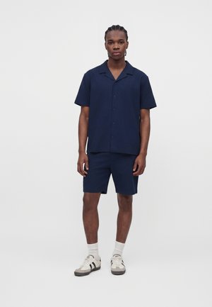 Mand stående iført en marineblå kortærmet knap-skjorte og matchende shorts med hvide sokker og beige sneakers med sorte striber.