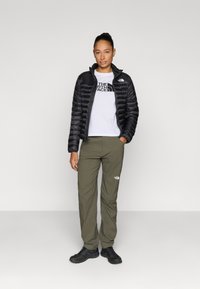 Μαύρο φουσκωτό μπουφάν, λευκό μπλουζάκι με λογότυπο, χακί παντελόνι και μαύρα παπούτσια. Λουστικά υλικά, εφαρμοστή σχεδίαση. Απλό φόντο.