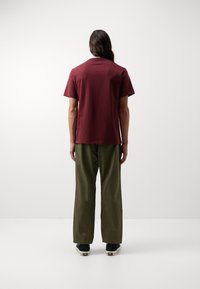 Camiseta de algodón burdeos de manga corta, combinada con pantalones sueltos de color verde oliva, mostrada desde la parte posterior contra un fondo claro.