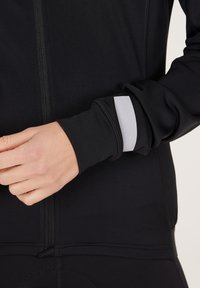 Zwarte sportjack met een getailleerd ontwerp. Getextureerde stof, zichtbare stiksels en een reflecterende zilveren accenten op de manchetten. Voorkant met ritsdetail.