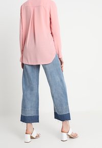 Ljusrosa långärmad blus med veckdetalj på baksidan, ihopparad med vida denimjeans med fransade fållar och blå accent nära botten.