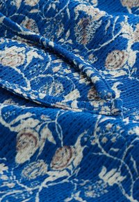 Tessuto blu texturizzato con motivi floreali in crema e rosa tenue. La trama è intricata con punti e dettagli visibili.