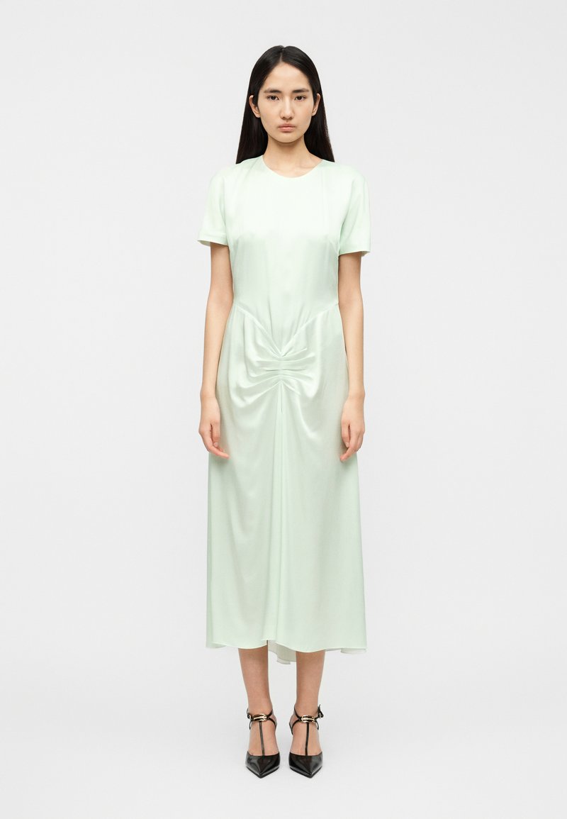 Midi φόρεμα σε μέντα πράσινο από απαλό ύφασμα, με κοντά μανίκια, στρογγυλή λαιμόκοψη και στριφογυρισμένη λεπτομέρεια μπροστά. Συνδυάζεται με μαύρες μυτερές γόβες.
