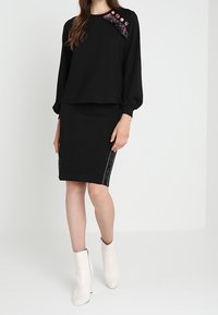 Svart långärmad topp med färgglada utsmyckningar på axlarna, tillsammans med en svart strukturerad pennkjol och vita ankelstövlar.