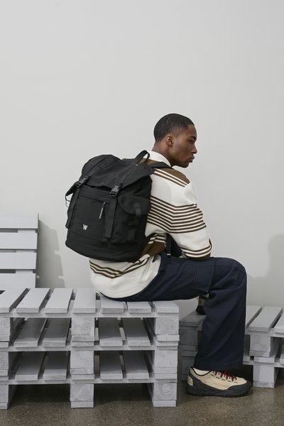 Sac à dos noir avec bretelles réglables, poche avant et porte-bouteille d'eau latéral. Le modèle porte un pull rayé et un pantalon bleu marine.