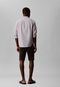 Άνδρας που στέκεται με την πλάτη γυρισμένη, φοράει ανοιχτόχρωμο μακρυμάνικο πουκάμισο με γυρισμένα μανίκια, μαύρα σορτς μέχρι τα γόνατα και μαύρα σανδάλια slide.