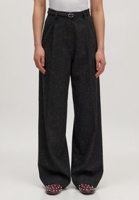 Vrouw in hoge taille, wijde donkergestippelde broek met een smalle zwarte riem, gecombineerd met gestudde bordeauxrode schoenen.
