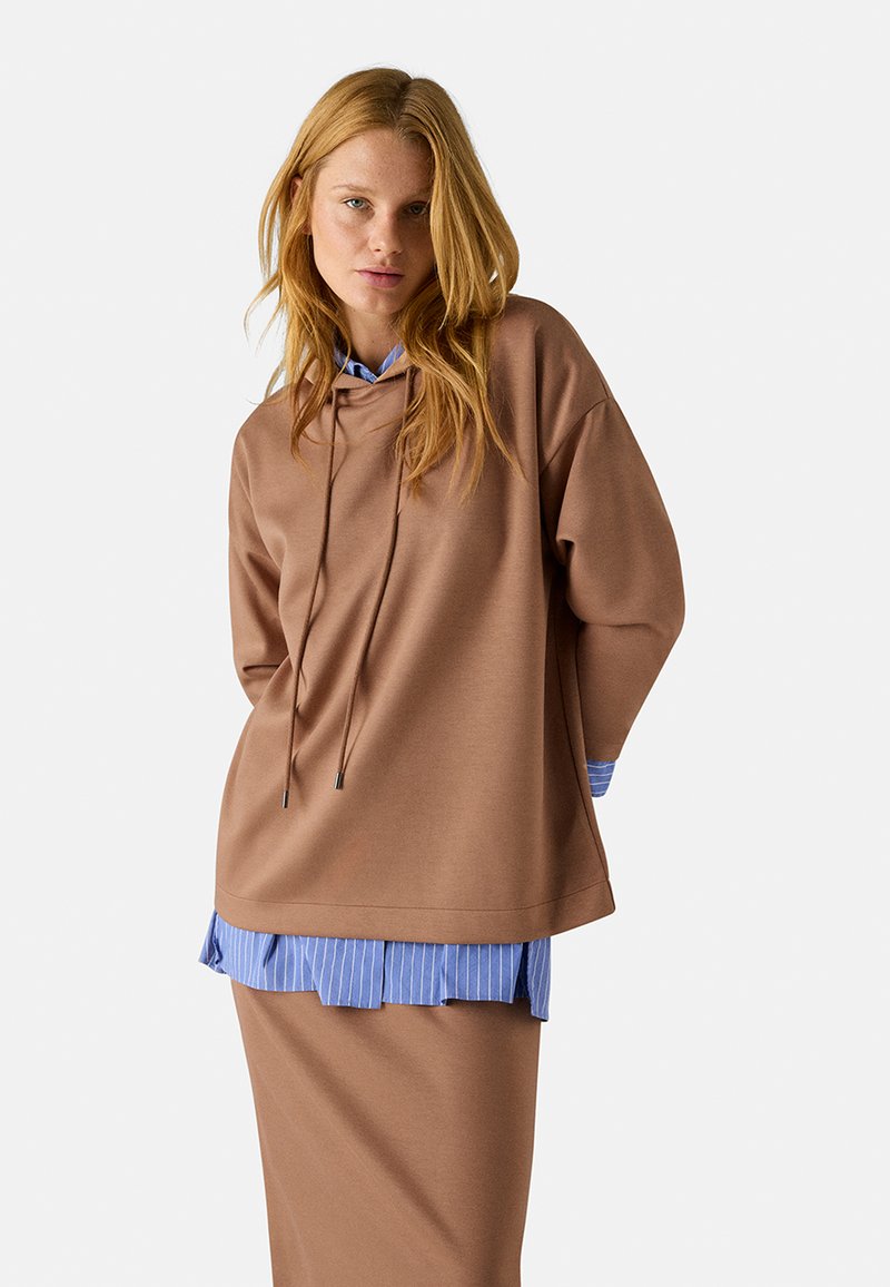 Sudadera oversized marrón con cordones sobre una camisa de rayas azul y blanca, combinada con una falda marrón a juego. Tejido suave y suave.