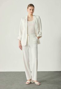 White tailored blazer over a white tank top, paired with white trousers and light sandals. Smooth fabric with a structured design, minimal detailing.