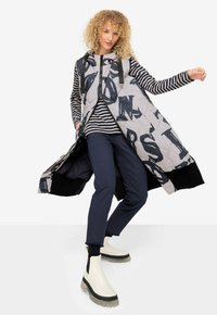 Chaleco acolchado largo en gris con patrones abstractos en negro, combinado con una camiseta a rayas en navy y blanco, junto con pantalones navy y botas blancas de tobillo.