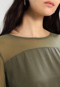 Top verde oliva con un collo arrotondato e una scollatura in rete trasparente, realizzato in un tessuto liscio, con cuciture a vista lungo i bordi.