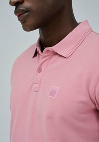 Polo de color rosa claro hecho de algodón, con cuello clásico, una tapeta de botones en la parte frontal y un logo bordado en el pecho.