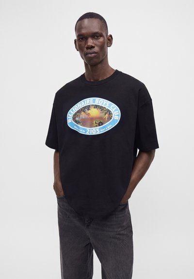 Man wearing a black oversized t-shirt with a blue oval logo reading "Billionaire Boys Club" and a sunset beach graphic, paired with dark jeans.
