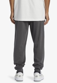 Person wearing dark gray jogger pants, white sneakers with "07" on heels, and a loose white shirt, shown from behind against a white background.