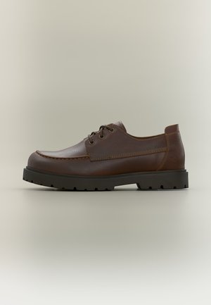 Chaussure en cuir marron à lacets avec une semelle épaisse en caoutchouc noir et des coutures détaillées, vue de côté sur un fond neutre.