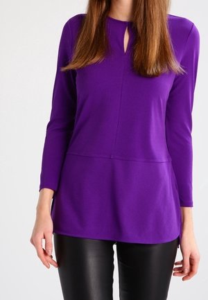 Vrouw die een paars shirt met lange mouwen en een klein sleutelgat bij de hals draagt, gecombineerd met zwarte leren broek, tegen een witte achtergrond.