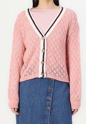 Kvinde iført en lyserød strikket cardigan med hvide og sorte kanter over en lyserød top, kombineret med et blåt denimskørt med knapper.
