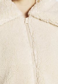 Giacca in pile color crema con chiusura a zip e colletto ampio. La texture è morbida, con un aspetto soffice e un design semplice.