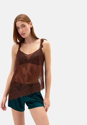 Jonge vrouw die een bruin kanten mouwloos topje draagt met gestrikte schouderbanden en donkergroene satijnen korte broek, staand tegen een effen lichte achtergrond.