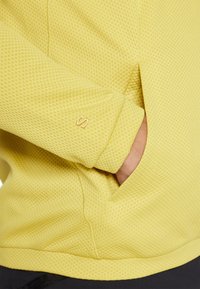 Pulover din plasă galben, cu un model texturat, având o buzunare și un mic logo brodat pe manșetă, punând în evidență un design ajustat.