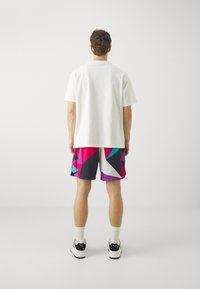 Camiseta blanca de algodón, combinada con pantalones cortos de malla multicolores con patrones geométricos en rosa, púrpura, verde azulado y negro. Zapatillas negras.