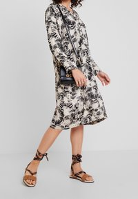 Sukienka w kwiaty w kolorze czarnym i kremowym z długimi rękawami, długości do kolan i dekoltem w kształcie litery V, w połączeniu z brązowymi zamszowymi sandałami i czarną torebką na ramię.