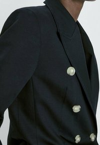 Blazer noir croisé en tissu lisse, doté de larges revers, de boutons argentés et d'une silhouette cintrée.