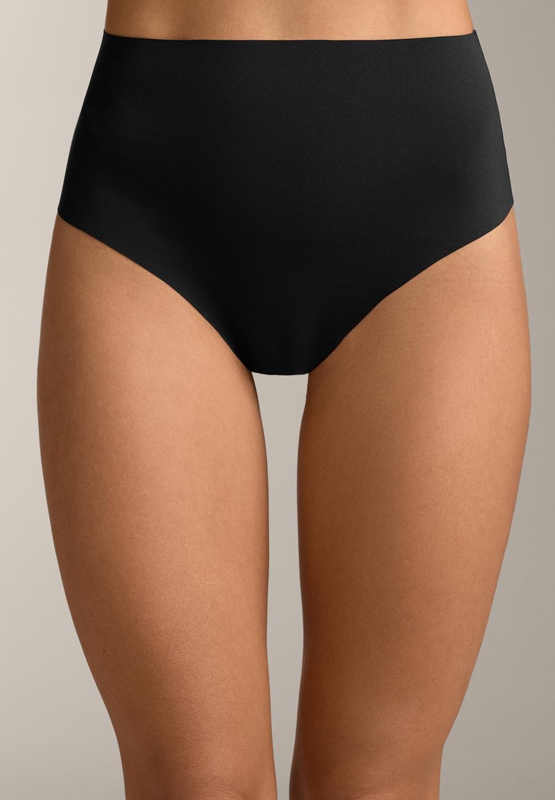 Femeie purtând lenjerie intimă neagră fără cusături, cu talie înaltă, stând în picioare pe un fundal neutru, arătând porțiunea mediană a corpului și partea superioară a coapselor.