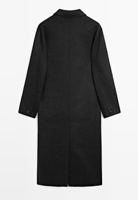 Cappotto lungo nero con colletto, polsini con bottoni e uno spacco centrale sul retro. Realizzato in un tessuto testurizzato, presenta un design minimalista.