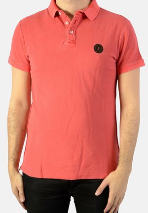 Koraalrode polo met korte mouwen, een gestructureerde kraag, een drieknoopsluiting en een cirkelvormig logo op de borst. Draagt bij donkere jeans.