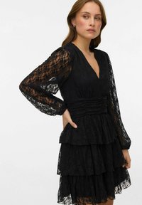 Femme portant une robe noire en dentelle avec des manches longues en tissu transparent, une jupe à volants et une taille froncée, debout avec une main sur la hanche.