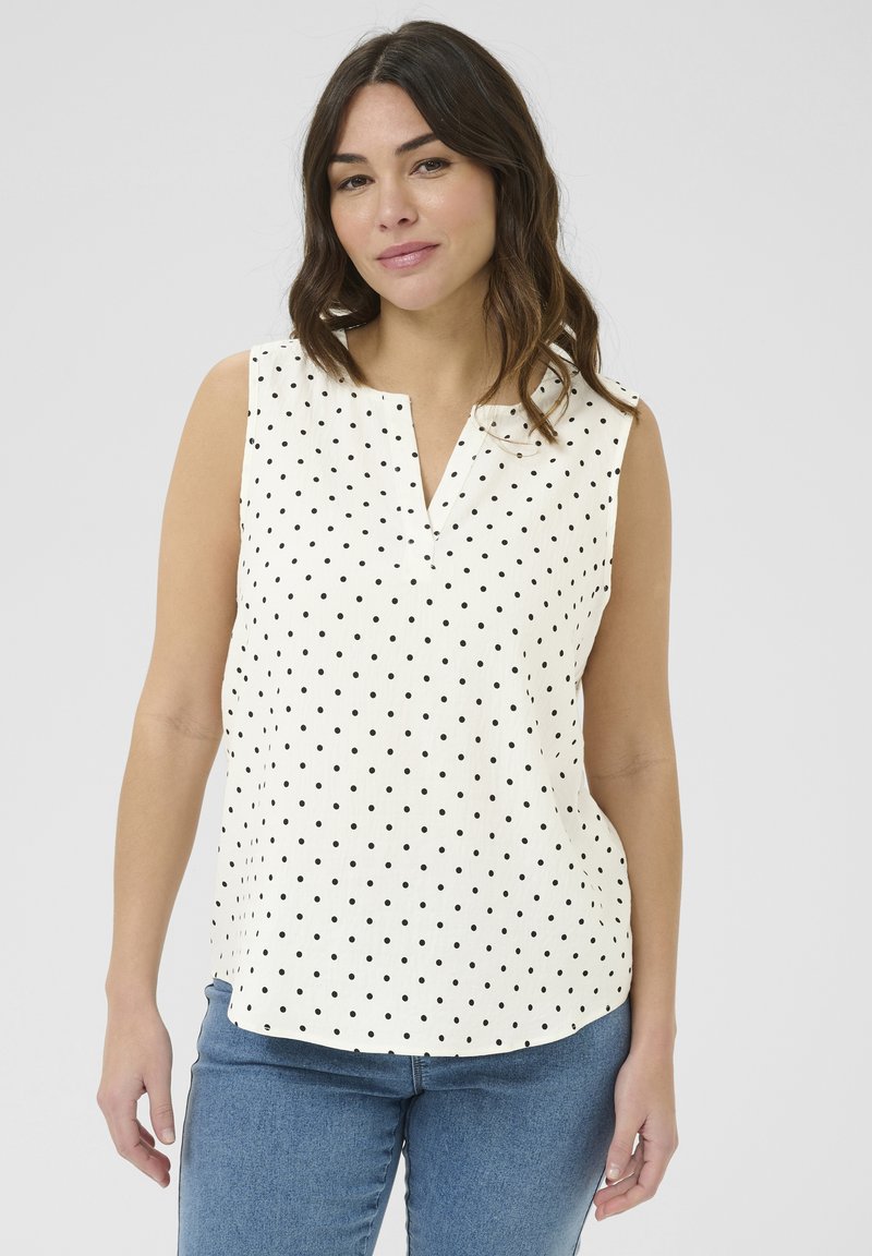Blouse sans manches en crème avec des pois noirs, présentant un décolleté en V et une coupe décontractée, fabriquée en tissu léger avec une texture lisse.