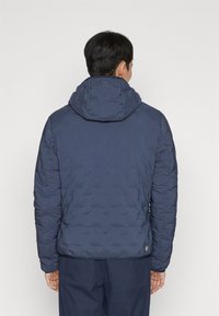 Man staat met zijn rug naar de camera, draagt een marineblauwe doorgestikte jas met capuchon en donkerblauwe broek, tegen een effen achtergrond.