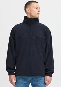 Pullover di pile navy con colletto alto, zip frontale a metà e tasca sul petto. Presenta polsini elasticizzati e una texture liscia.