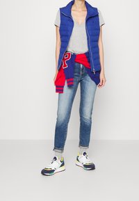 Chaleco azul acolchado, camiseta gris de cuello en V, suéter rojo atado a la cintura, jeans azules desgastados y zapatillas multicolores. Atuendo casual.