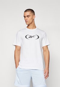 Hvit bomull T-skjorte med en svart logo i midten, kortermet og rund hals. Brukt med lyseblå perforerte shorts.