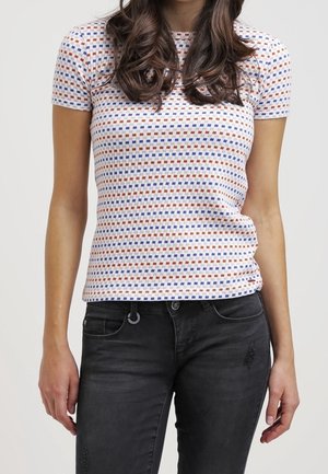 Woman wearing a short-sleeve white top with blue and brown square patterns and black denim jeans against a plain background.