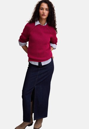 Donna con capelli ricci che indossa un maglione rosso sopra una camicia a righe, gonna lunga in denim scuro con spacco frontale e stivali marroni su sfondo bianco.