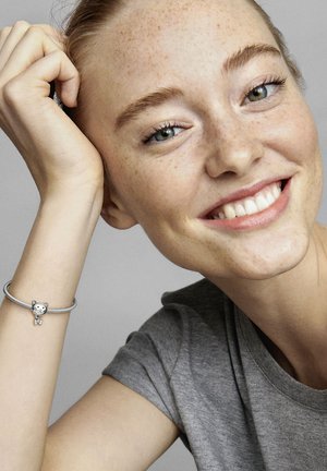Bracciale in argento con un charm a forma di gatto che presenta un sorriso e un papillon, indossato sul polso di una persona dalla pelle chiara. Sfondo di una maglietta grigia.