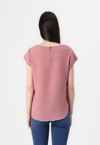 Top rosa a maniche corte in tessuto testurizzato con orlo arrotondato. Presenta una cerniera sul retro, abbinato a jeans di denim blu.