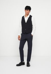 Bărbat îmbrăcat într-un vest bleumarin, cămașă albă, pantaloni bleumarin asortați și pantofi negri de haine, stând pe un fundal alb simplu.