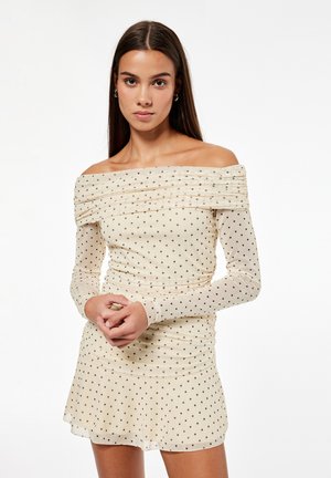 Junge Frau mit langen braunen Haaren, die ein schulterfreies beiges Kleid mit schwarzen Punkten trägt und mit verschränkten Händen da steht.