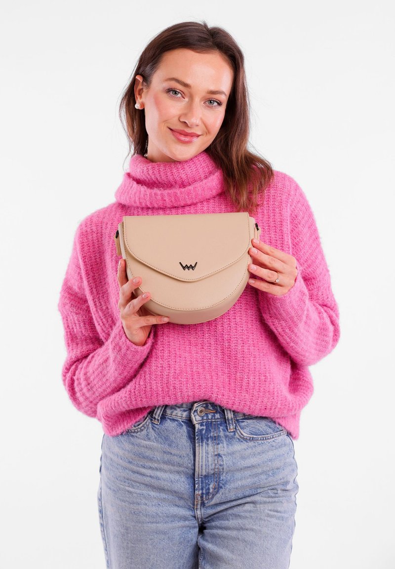 Béžová, polokruhová kabelka s klopou a embosovaným logem. Hladká kožená textura. Modelka drží kabelku na bílém pozadí.