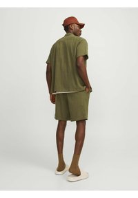 Olijfgroene korte mouwen knoopshirt en bijpassende shorts, gemaakt van gestructureerde stof. Bruine sokken en lichte instapsandalen.