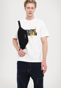 Black sling bag with a zip closure, worn on the shoulder, paired with a white T-shirt featuring yellow graphic text and navy blue pants.