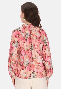 Bluză florală cu un model multicolor în nuanțe de roz, piersici și verde. Mâneci lungi cu un guler plisat și material moale.