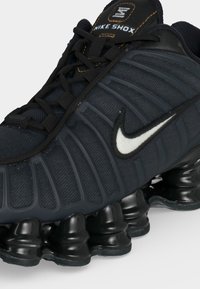 Μαύρο sneaker Nike Shox με υφή στην επάνω επιφάνεια, λευκό λογότυπο Nike και χαρακτηριστική διαχωρισμένη μαύρη επενδυμένη σόλα.