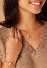 Gouden ketting en armband met een bloemenhanger bezet met heldere stenen. Model draagt een beige gebreide trui en laat de sieraden op haar hals en pols zien.